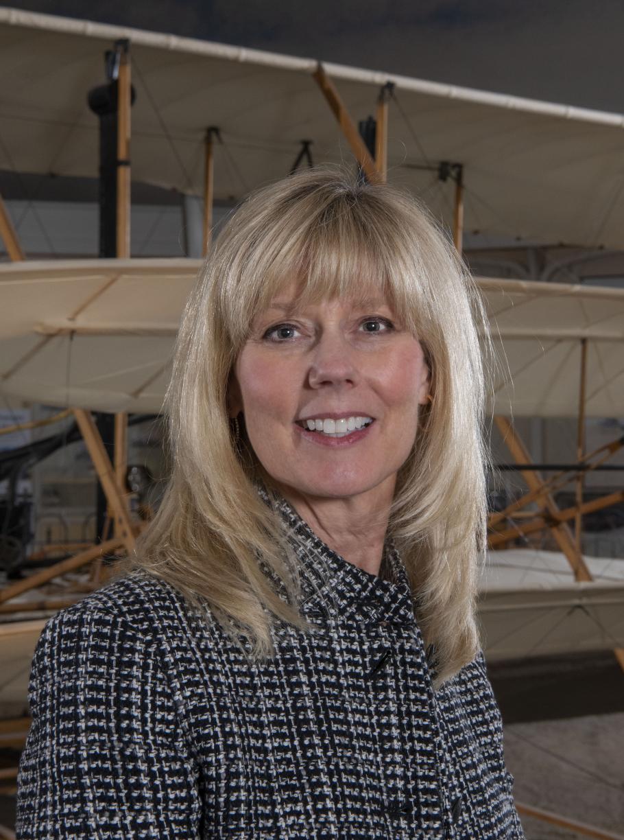 A blonde woman smiles at the camera with a plane behind her. 