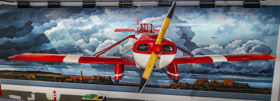 A red and white airplane hang in front of a mural of a Western landscape.