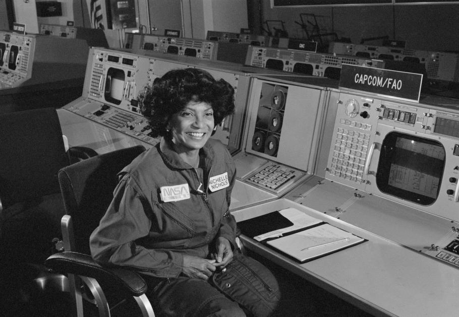 A woman, Nichelle Nichols sitting and smiling in a room with large desks and computers.