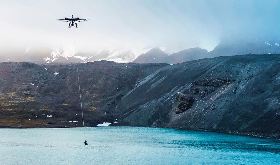 A hexacopter drone hovers above a lake in Antarctica. A long cord attached to the drone is fitted with a device to gather water samples from the lake, with minimal contamination.