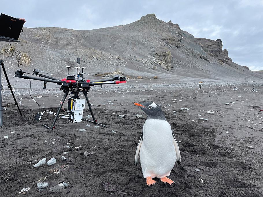 A drone sits on barren grey soil in Antarctica, while a penguin stands in the foreground, his head cocked to his right as he looks at his surroundings.