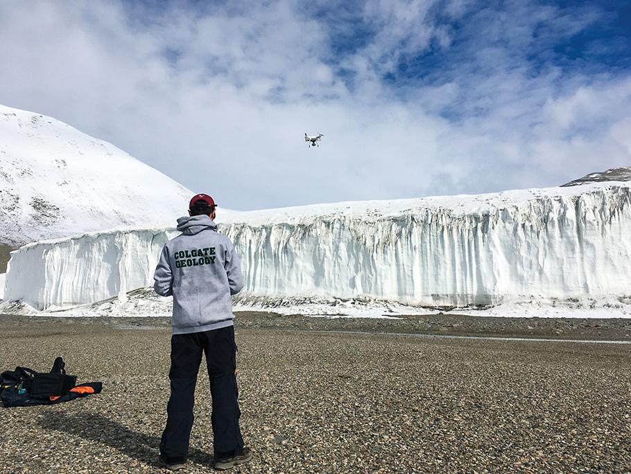 Geologist Joe Levy stands in front of a massive glacier jutting out of the surface of Antarctica, flying a drone via remote control.