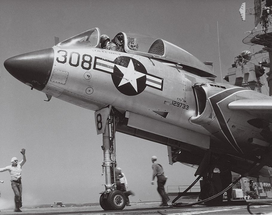 Two sailors are dwarfed by an F7U on the deck of an aircraft carrier. A distinguishing characteristic of the F7U is its long nose-landing-gear strut.