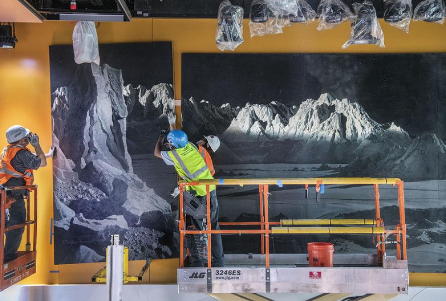 Two people in hard hats on a lift hold up a large panel and are in motion, placing the panel on the wall next to the other panels of the mural.