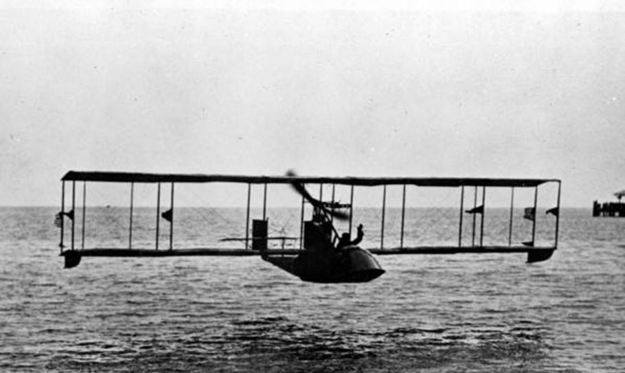 A black and white image of a plane flying over the water. 