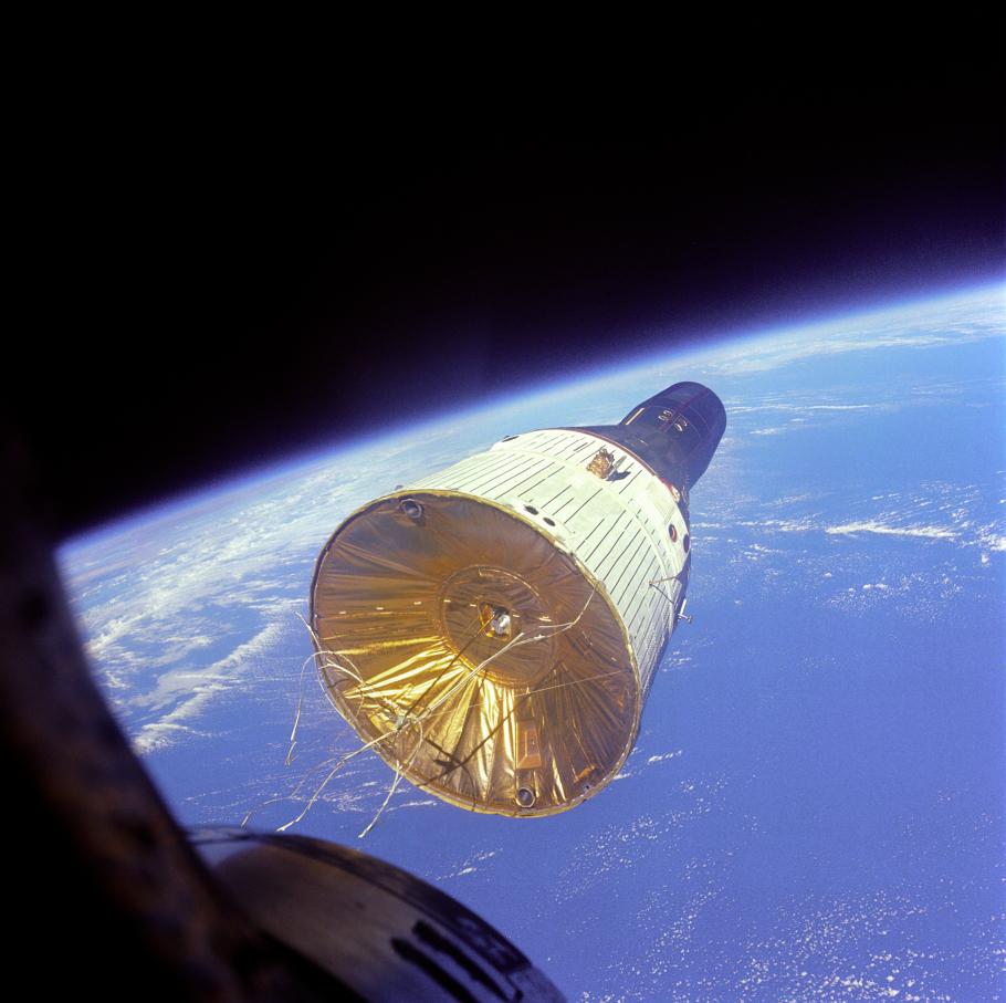 A three-quarters view of a conical shaped command module floating above the earth. The edge of the spacecraft taking the photo can be seen in the bottom lefthand corner.
