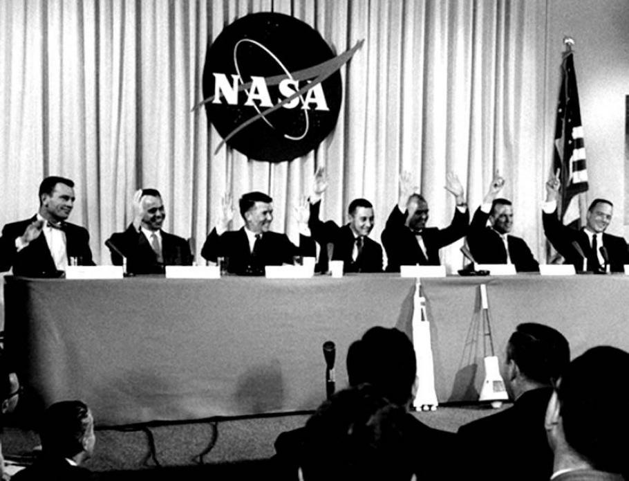 Seven men sit behind a table in front of an audience, each with name cards and microphones in front of them. All seven are raising their hands. 