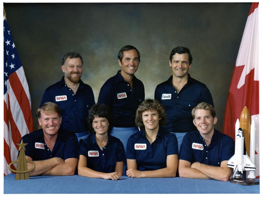Astronaut group portrait wearing navy polo shirts with NASA logo on right breast--three men standing behind two men with two women seated between them.  American flag on pole to the left, NASA flag on pole to the right. On the table: replica of gold astronaut pin on left, model of the space shuttle on the right.