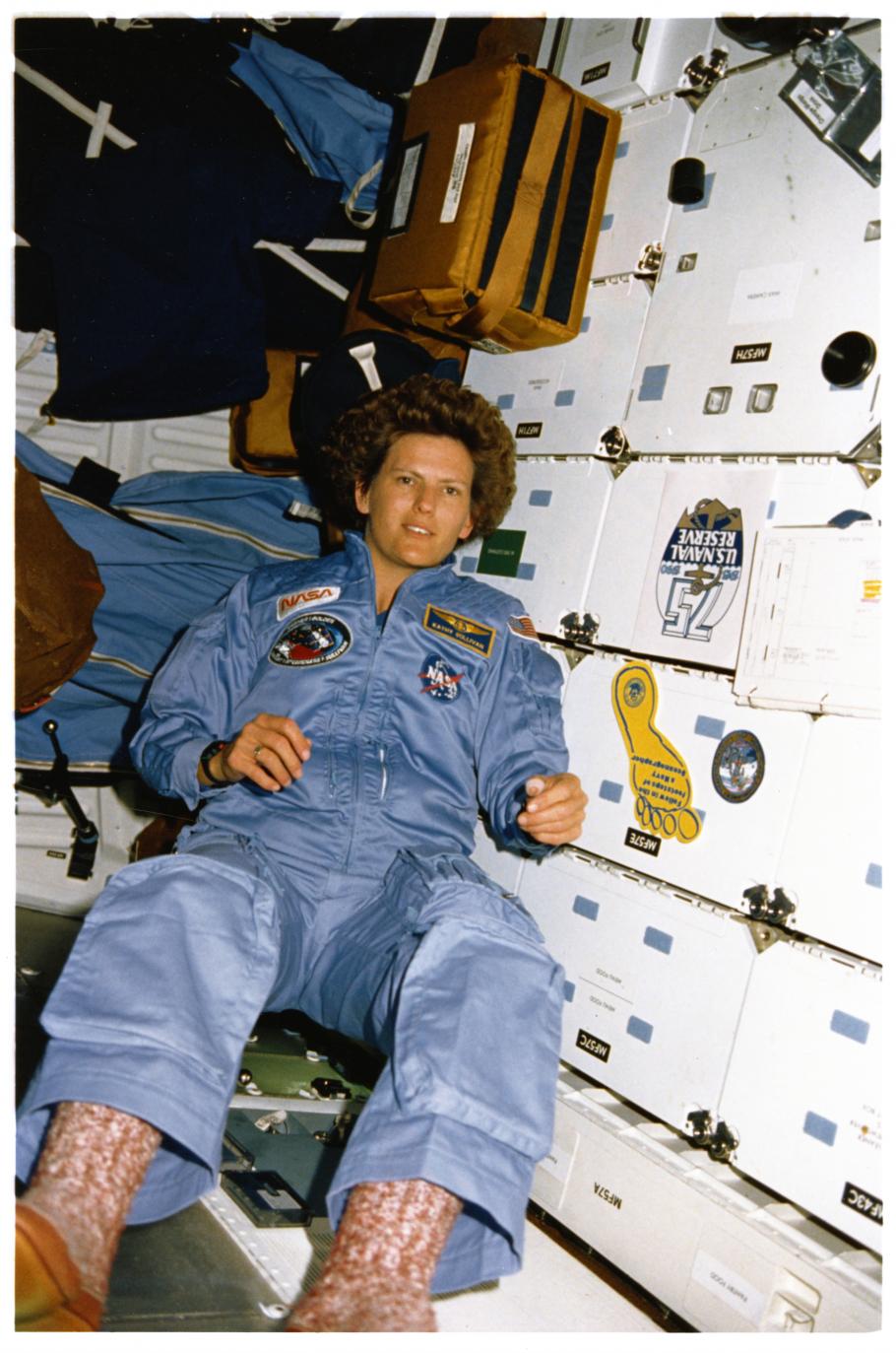 Woman in a light blue NASA flight suit floats weightlessly in front of the white lockered walls of the Space Shuttle.