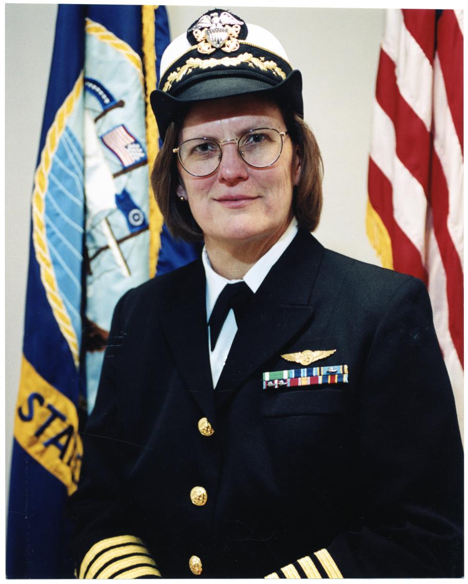 Portrait of a woman wearing glasses in a naval uniform with yellow stripes at the ends of the sleeves.  Her cap has a white top and black brim with a Navy insignia.  Behind her are the Navy flag and US flag.