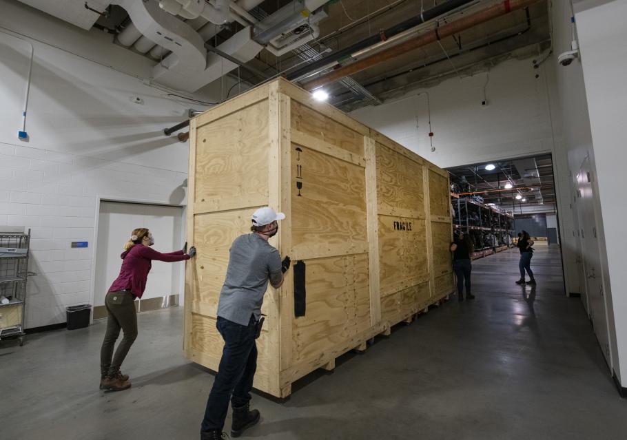 Staff members push a large wooden crate through a large hangar setting.