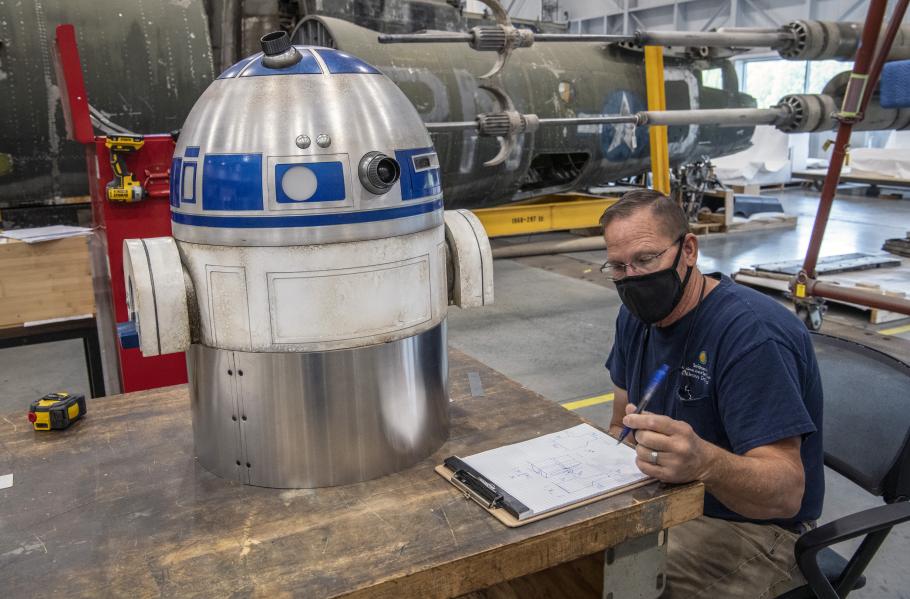 Man sits at a table writing on a pad of paper next to the top half of a model of R2-D2, a silver and blue droid.