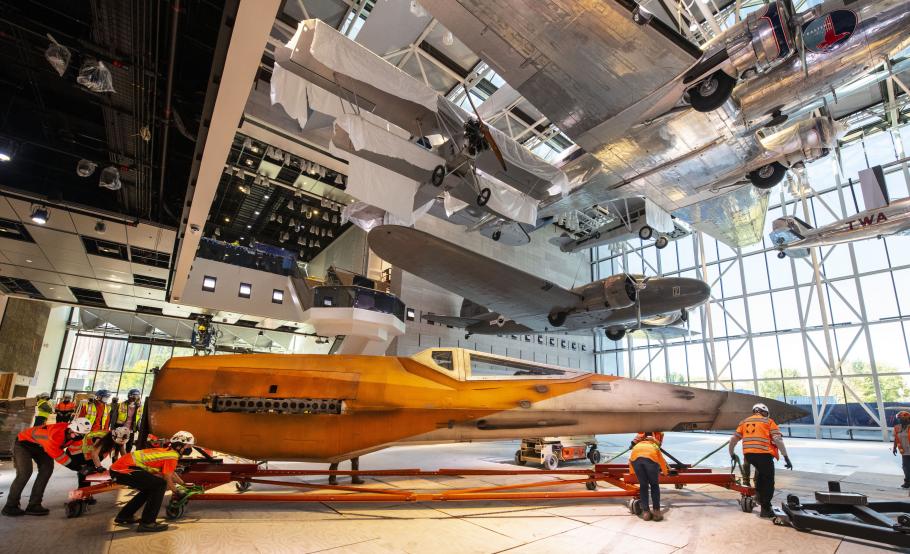 A group of workers in construction vests and hats move the fuselage of the X-wng, a pointed orange and white spacecraft, through a nearly-empty gallery with aircraft hanging above.