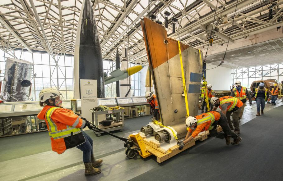 A group of people in orange construction vest and hat push the wing of an X-wing down the hall of a museum with rockets in the background.