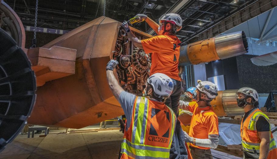 Four workers in construction vests and helmets assemble the engines of a Star War X-wing, an orange flying vehicle.