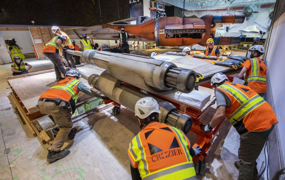 A group of workers in orange construction vests and helmets move part of the X-wing in the construction area at the Museum in DC.