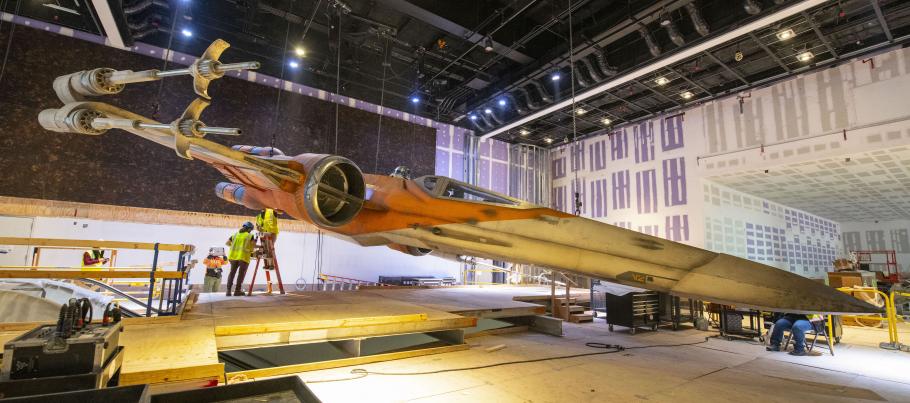 A cream an orange full-size X-wing hanging a few feet over the ground as it is raised.