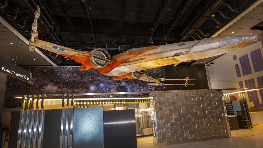 X-wing, a weathered cream and orange space vehicle hangs over an unstocked store and ticket area.