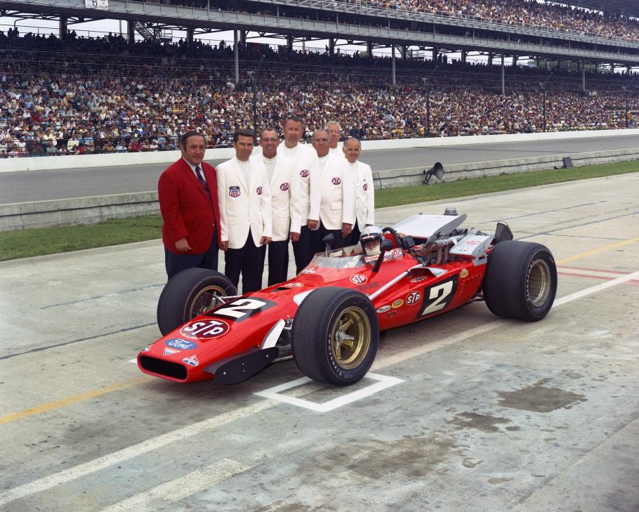 One man in a red jacket and six med in white jackets standing behind a red race car.