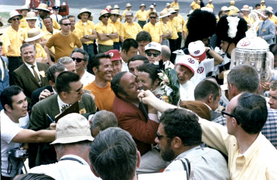 Two men are surrounding by a crowd. Andy Granatelli places a kiss on Mario Andretti's cheek.