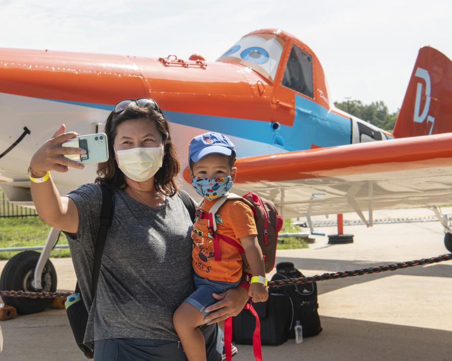 Woman holding a small child takes a selfie in front of a cartoony-looking aircraft.