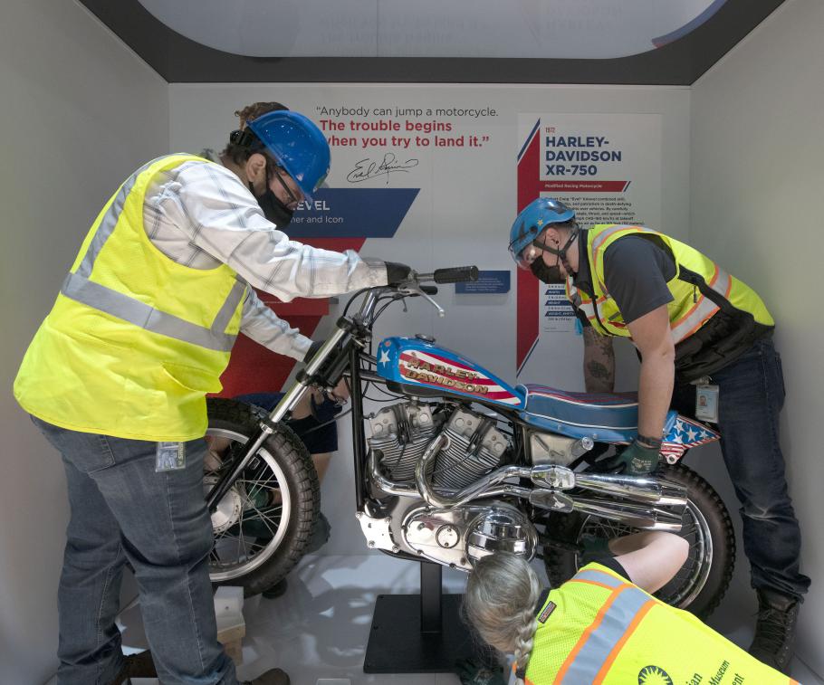 Two people in yellow construction vests and helmets install a motorcycle in a display case.