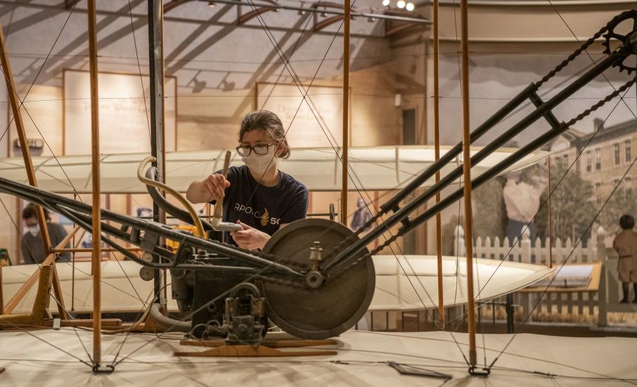 A woman wearing a face mask and an Apollo 50 t-shirt uses a brush to clean the 1903 Wright Flyer
