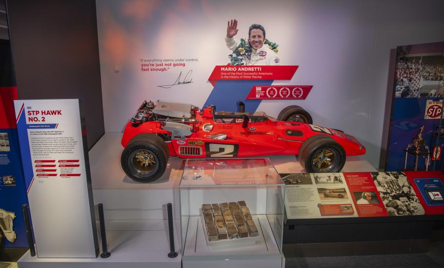 Red race car on display with photo of Mario Andretti on the wall behind it.