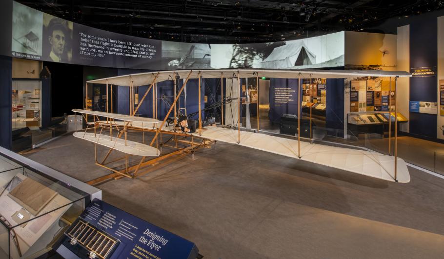 A view of the Wright Flyer in situ, with images and text on the wall behind it. 