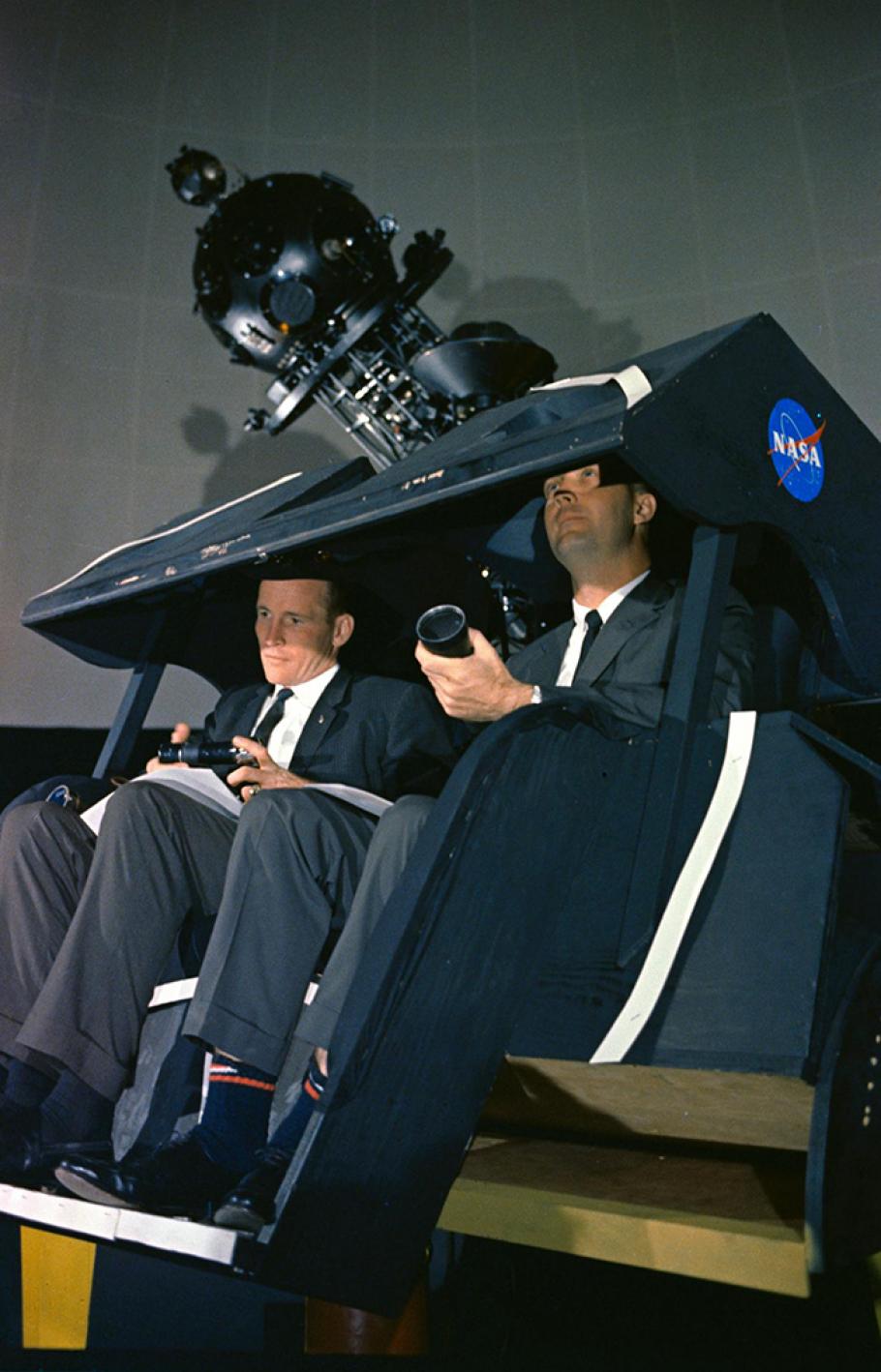 Two men sitting in a simulation training device that is enclosed overhead but open in the front