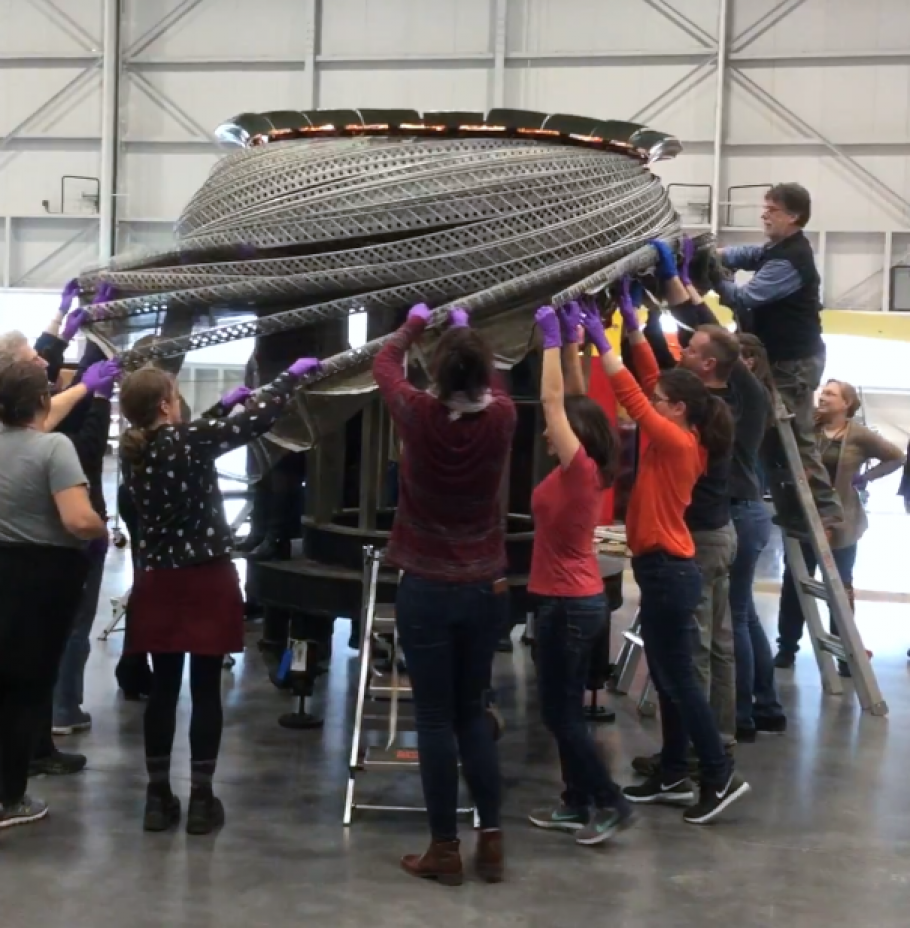 A group of approximately 10 individuals lifting a large object above their heads in a open interior space.