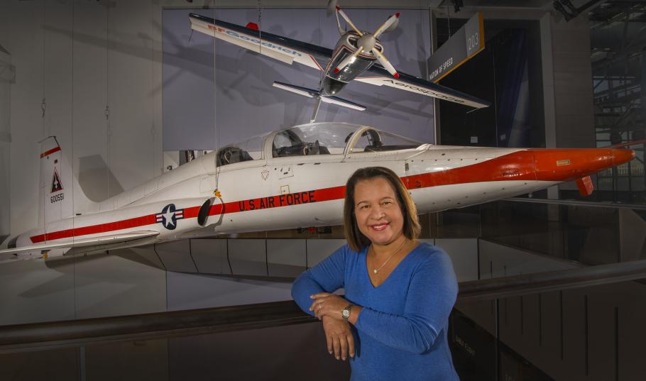 A woman smiles at the camera. Behind her is an orange and white airplane. 