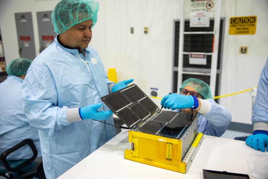 Two men in clean-room robes, gloves, and head coverings work on a CubeSat, a small satellite.