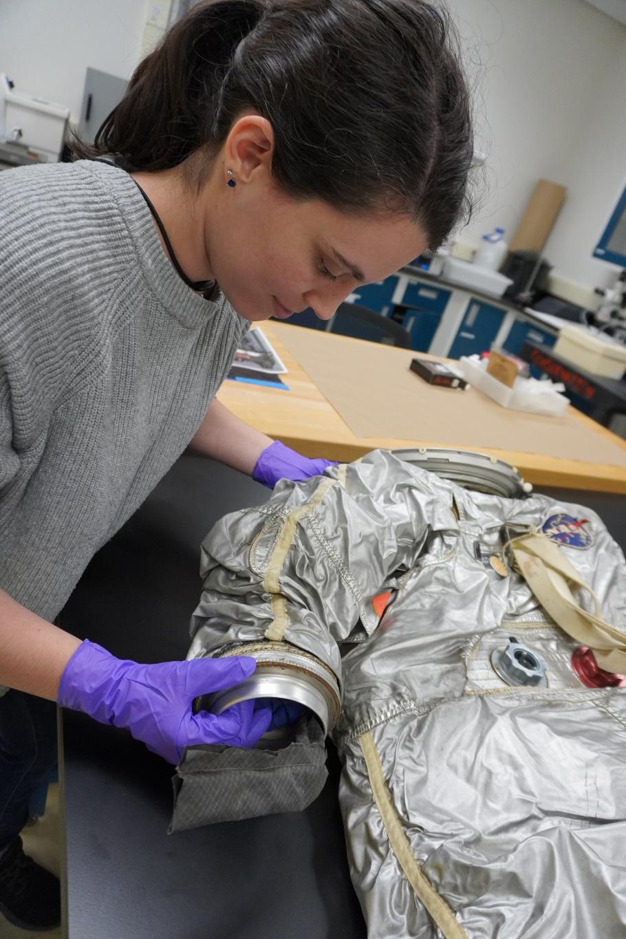 A woman wearing purple gloves holds a silver spacesuit. 