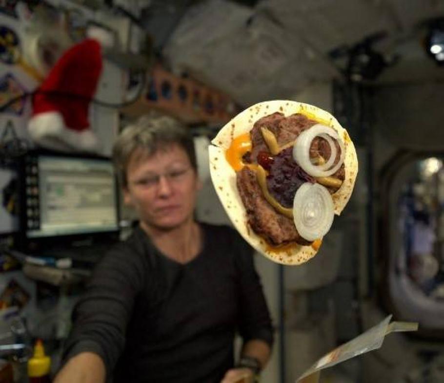 In the foreground, a tortilla with meat, cheese, and onions floats in zero gravity. A woman is in the middle ground. In the background are the interior walls of the International Space Station. 