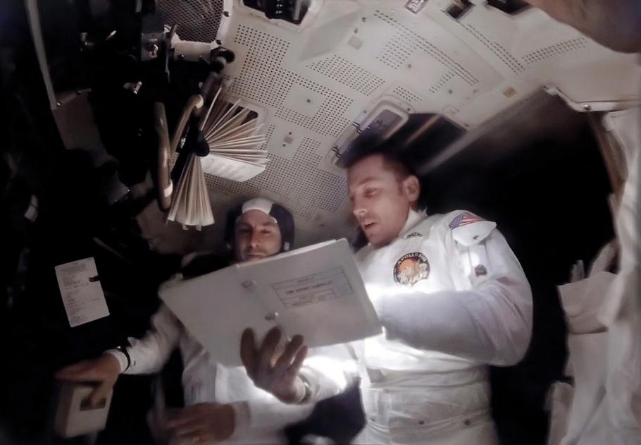 Astronauts Jim Lovell and Jack Swigert stand next to one another in the cramped Apollo 13 control module, reviewing a large booklet titled  "CSM Entry Checklist,” which contained the procedures necessary for re-entry. 