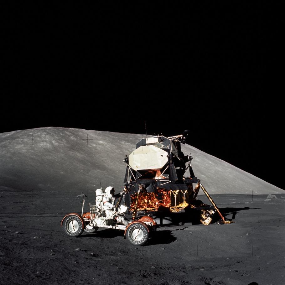 A lunar module and a lunar roving vehicle is photographed on the surface of the moon. An astronauts is sitting in the lunar roving vehicle.