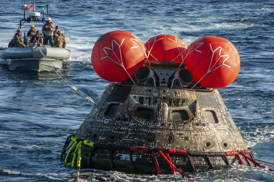 A space capsule is seen in the ocean in the foreground and a lifeboat with people in the background.
