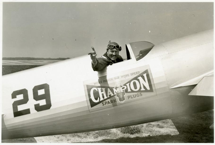 Side view of Roscoe Turner seated in the cockpit of his LTR-14 "Meteor" airplane on the ground. Turner points his right hand and winks.