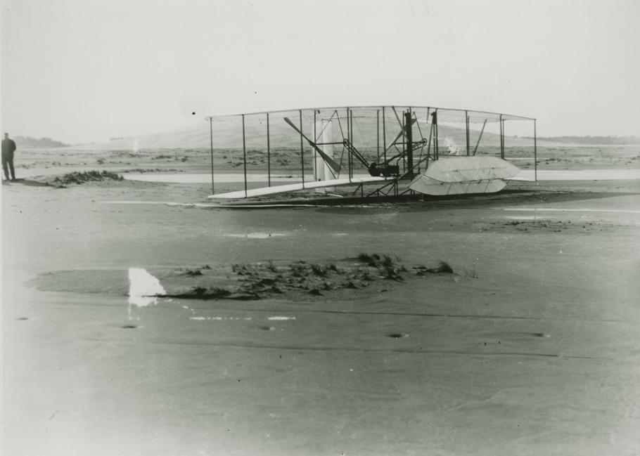 A black and white photo of a biplane on a sandy beach with a man standing the the far left. The front elevator is almost perpendicular to the plane as it is damaged.
