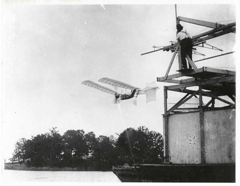 One-half left rear view of a Langley Aerodrome No. 5 in flight over the Potomac River just after launch. A man watches from a houseboat at top right.