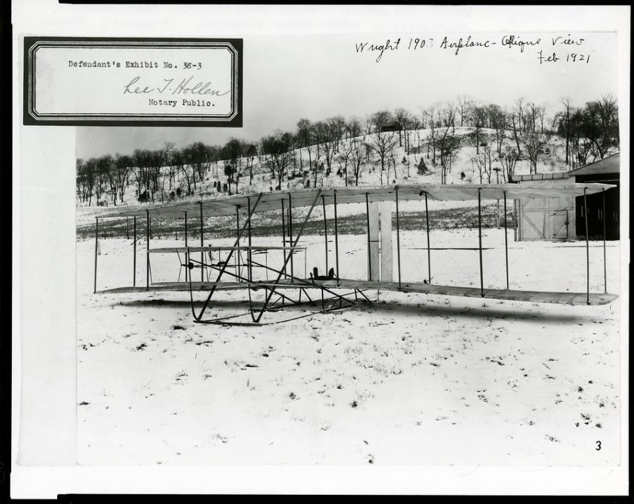 The Wright Flyer in the foreground of a snowy field, seen from the front. There is an exhibit number stamped at the top left.