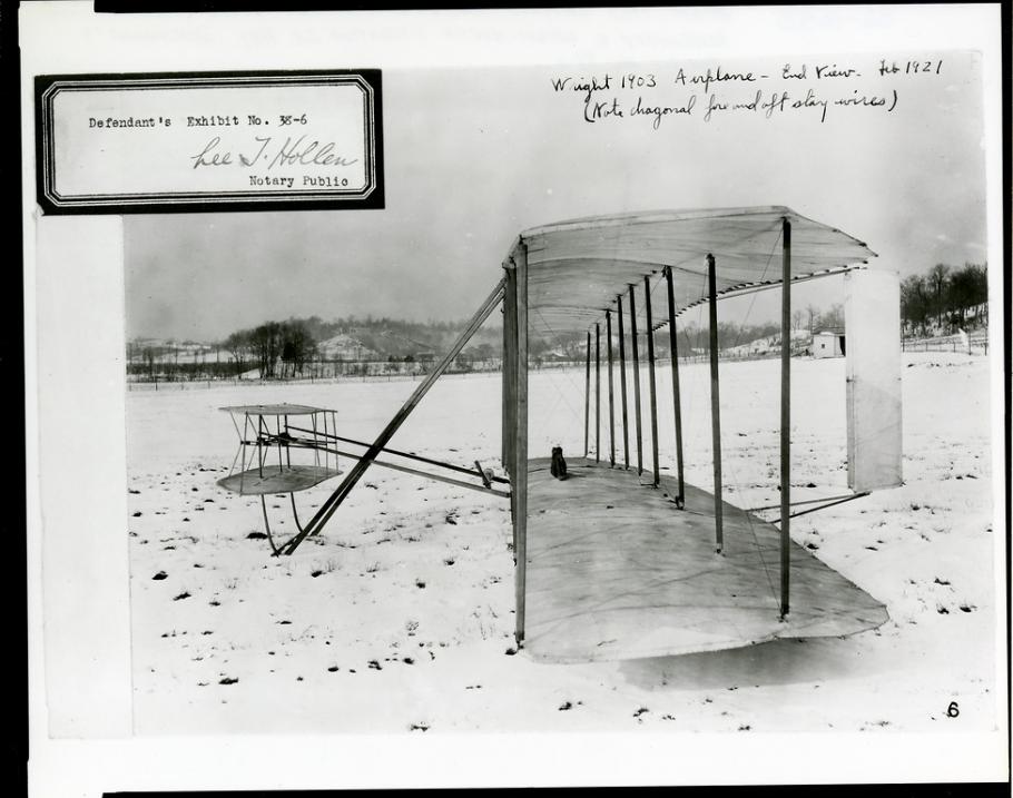 The Wright Flyer as seen from the side in a snowy field. There is an exhibit number stamped at the top left.