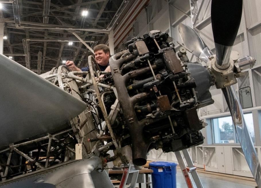 A man stands on a ladder by the nose of an aircraft with its internal mechanics exposed.