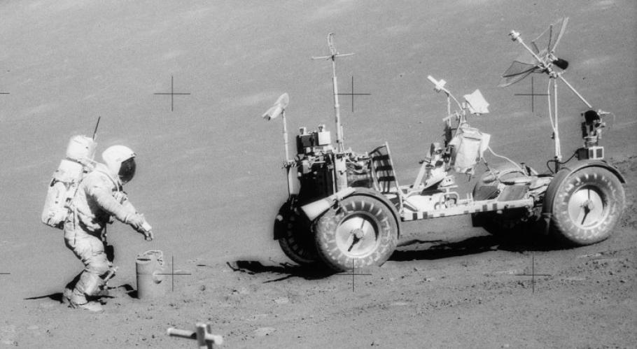 On the Moon, an astronaut in a space suit seems to set down or pick up a bucket by the Lunar Roving Vehicle. 