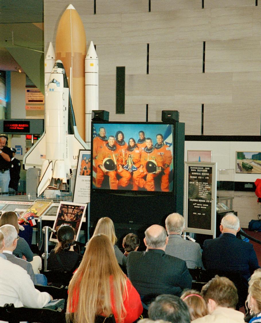 Astronauts are shown on a small tv screen while a small ground looks towards it.