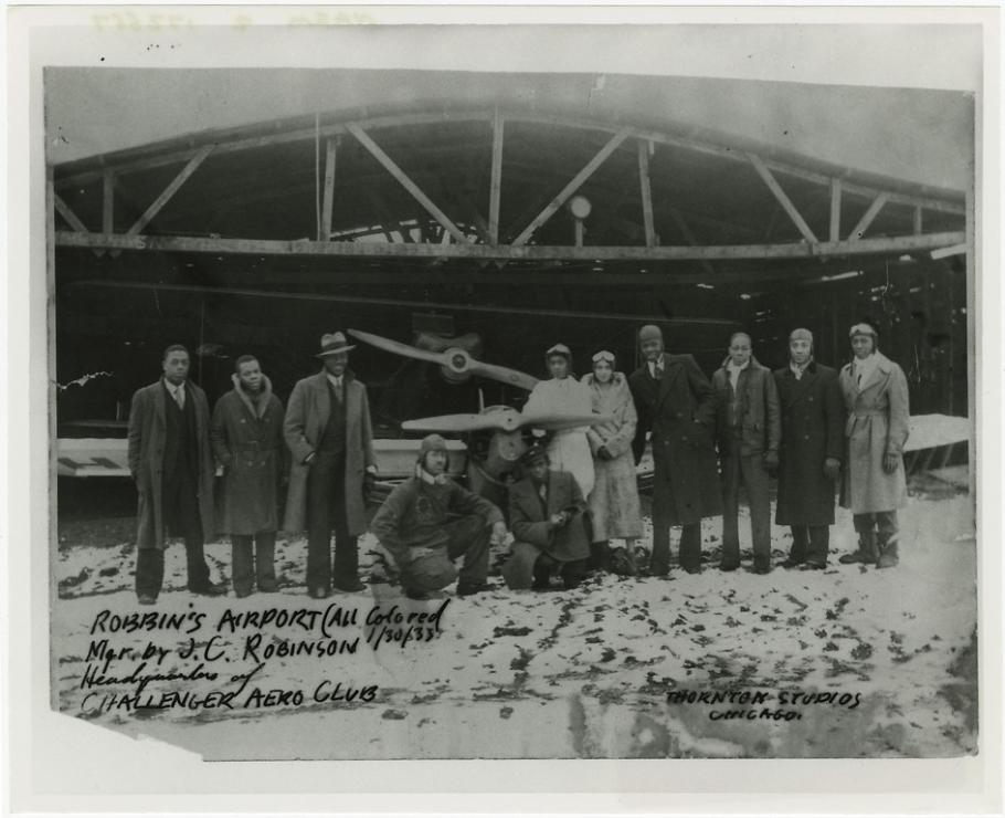 A group of eleven people stand in a line next to each other in front of an aircraft hangar. An airplane can be seen directly behind the group.