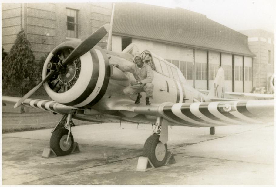 Sepia toned image of Jesse Leroy Brown squatting on the right side wing of an aircraft as he poses for a picture.