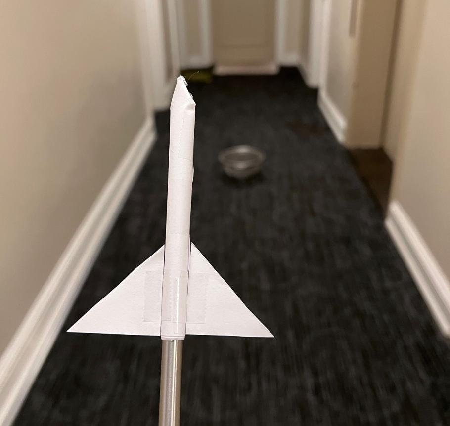 A paper rocket on a straw in the foreground. A colander on carpet in the background.