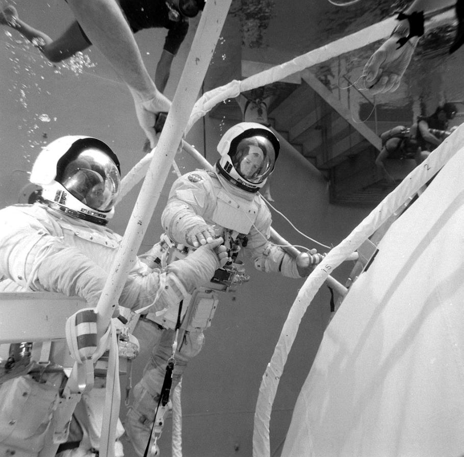 Black and white image of two astronauts attached to connected device underwater. They appear to be repairing a larger object.